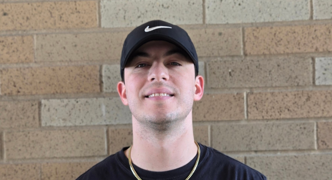 Jordan Schilling is pictured against a brick background, wearing a black Nike baseball cap, a gold chain, and a black t-shirt.